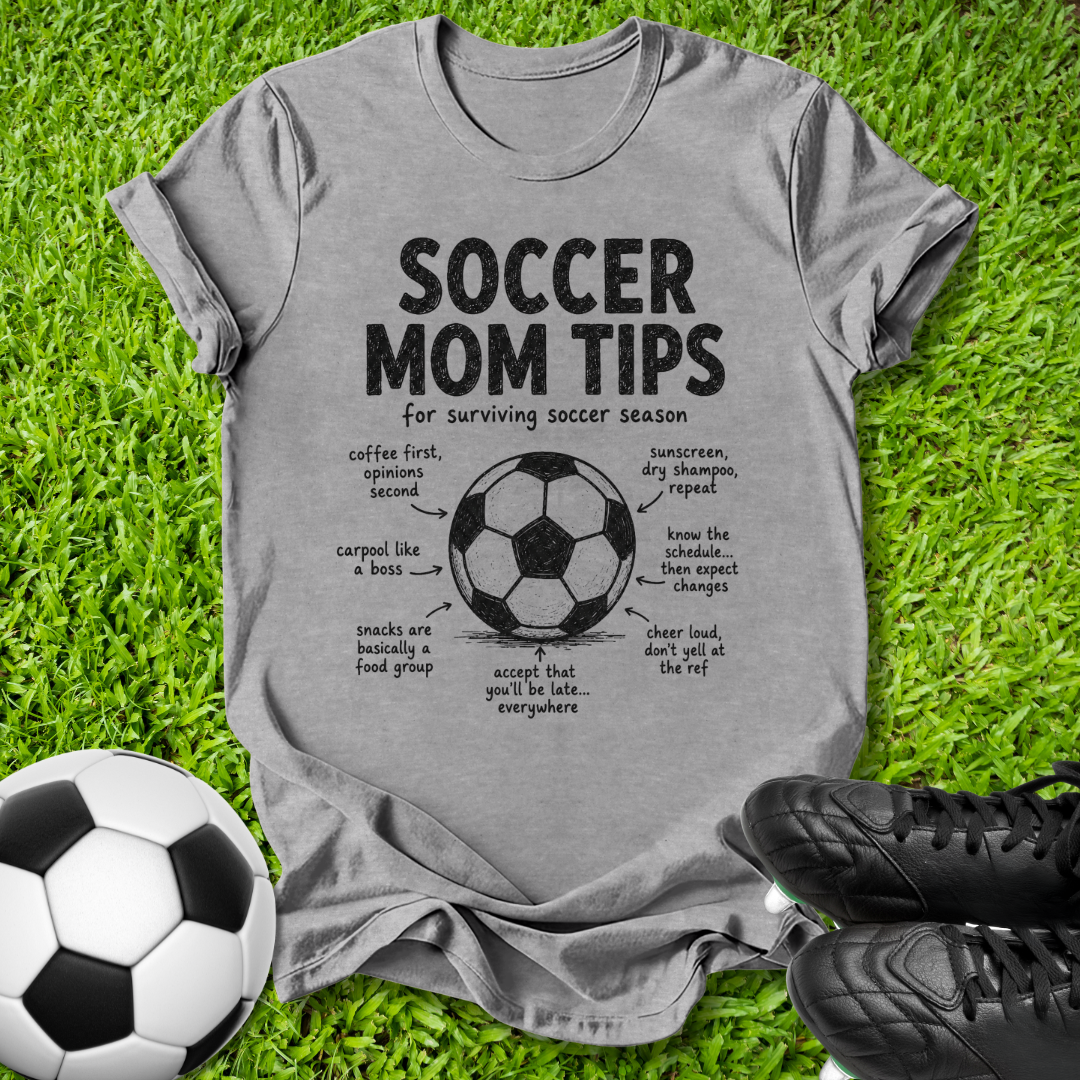 A heather gray graphic t-shirt laid flat on green grass, featuring a black and white soccer ball, black cleats, and the text 'SOCCER MOM TIPS' with a soccer diagram. The t-shirt is styled as a flat-lay with thematic props.