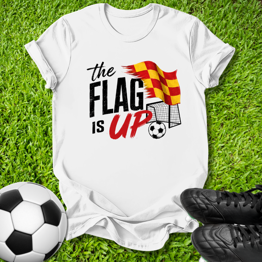 A white graphic t-shirt laid flat on green grass, featuring the text 'the FLAG IS UP' with a red and yellow striped flag, soccer ball, and goal illustration. A black-and-white soccer ball sits on the left, and black cleats are on the right.