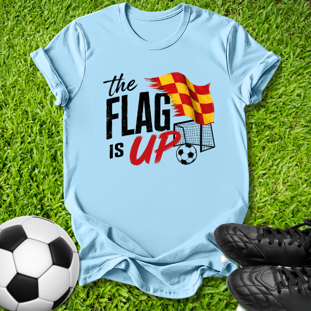 A light blue graphic t-shirt laid flat on green grass, featuring the text 'the FLAG IS UP' with a red and yellow striped flag and soccer ball illustration. A black-and-white soccer ball and black cleats are placed around it as props.