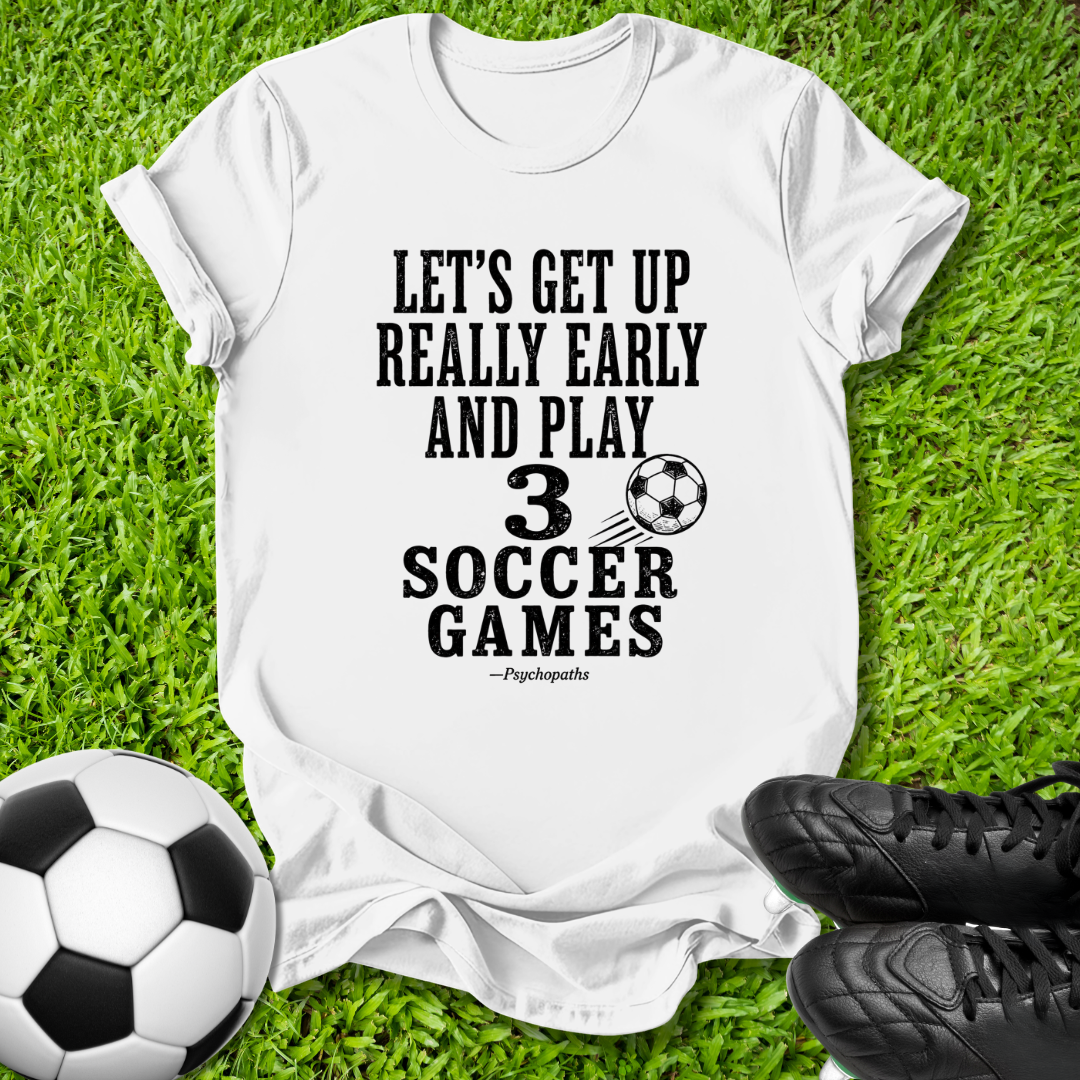 A white graphic t-shirt laid flat on green grass, featuring the black text 'LET'S GET UP REALLY EARLY AND PLAY 3 SOCCER GAMES' with a soccer ball graphic and the name 'Pssssshh'. A soccer ball and black cleats are placed around it as props.