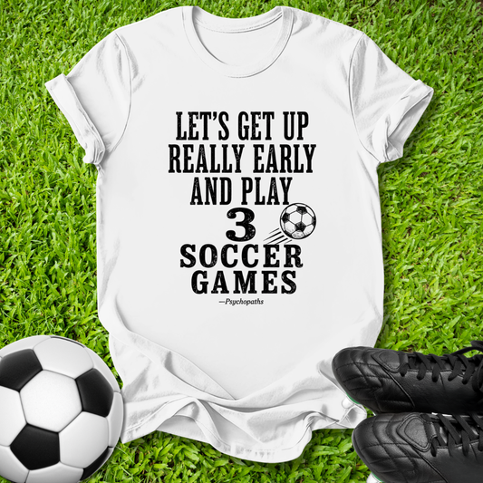 A white graphic t-shirt laid flat on green grass, featuring the black text 'LET'S GET UP REALLY EARLY AND PLAY 3 SOCCER GAMES' with a soccer ball graphic and the name 'Pssssshh'. A soccer ball and black cleats are placed around it as props.