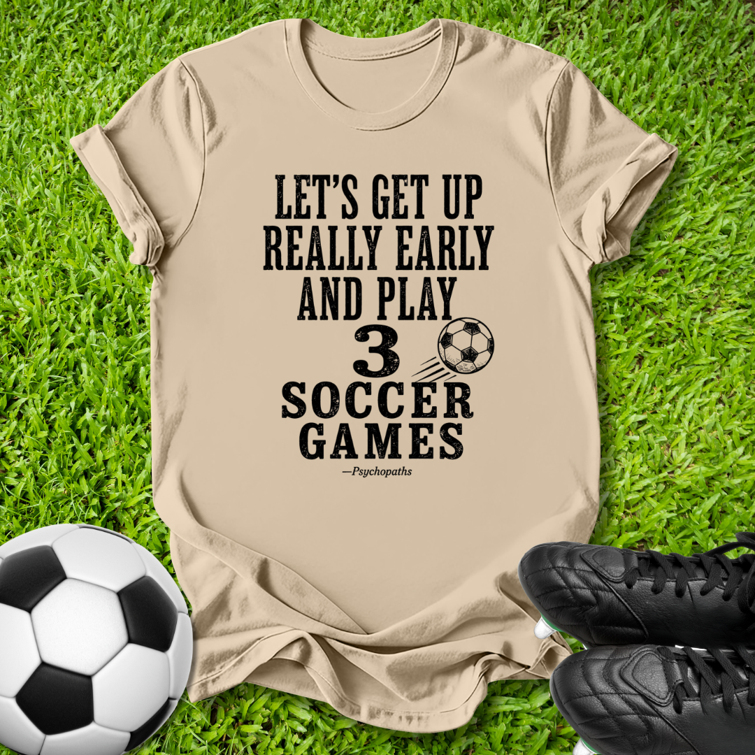 A beige graphic t-shirt laid flat on green grass, featuring the black text 'LET'S GET UP REALLY EARLY AND PLAY 3 SOCCER GAMES' with a soccer ball graphic. A soccer ball and black cleats are placed around it as props.