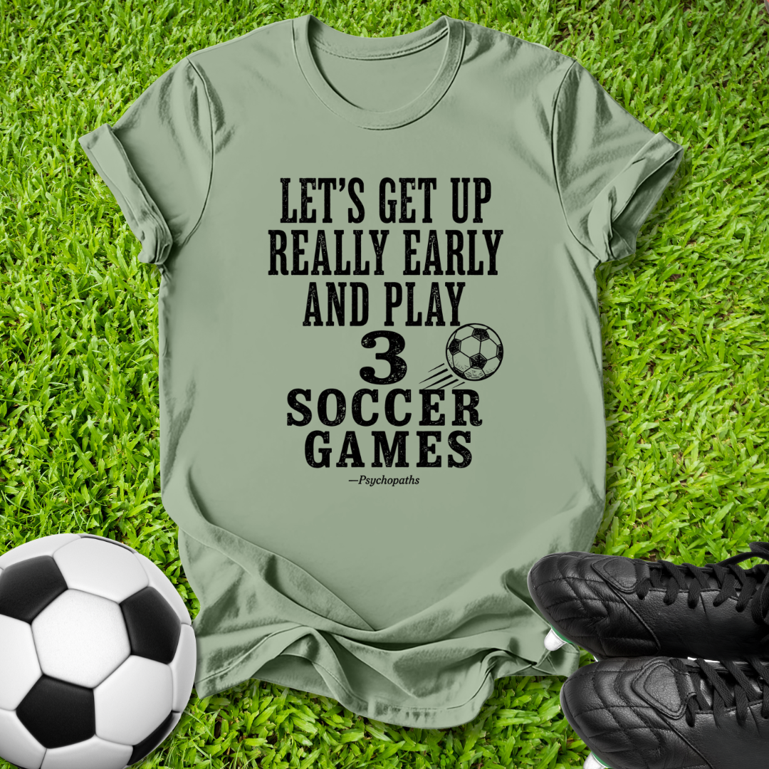 An olive green graphic t-shirt laid flat on green grass, featuring a black printed slogan about soccer. A soccer ball is visible in the bottom-left corner and black cleats are in the bottom-right, creating a themed outdoor flat-lay composition.