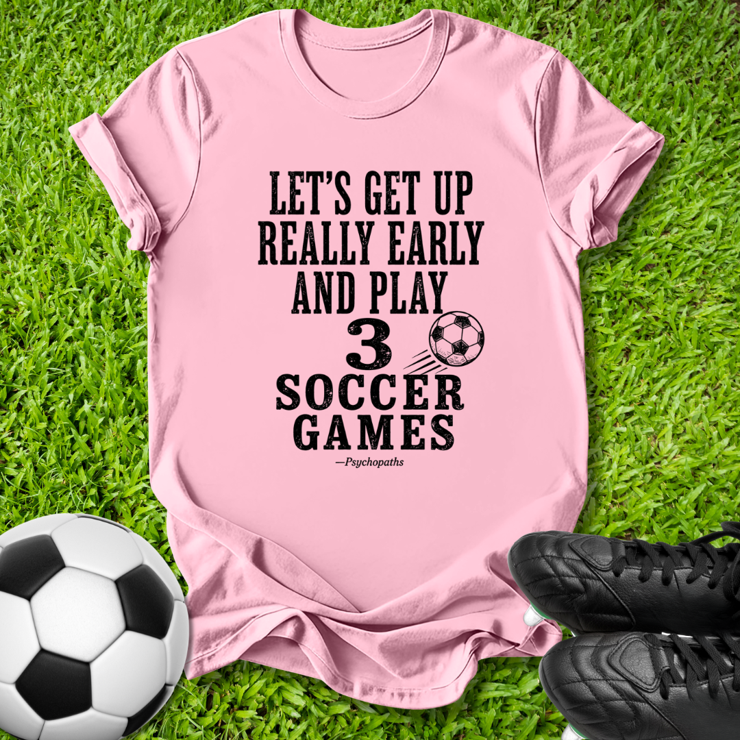 A pink short-sleeve t-shirt with a black soccer-themed text graphic reading 'LET'S GET UP REALLY EARLY AND PLAY 3 SOCCER GAMES' and a small soccer ball illustration, laid flat on green grass with a soccer ball and black cleats nearby.