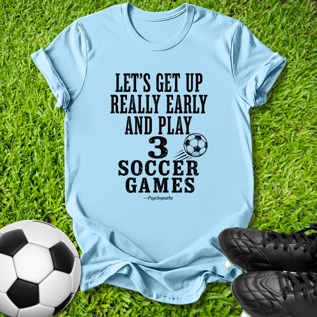 A light blue graphic t-shirt laid flat on green grass, featuring a black printed slogan about soccer. A soccer ball and black cleats are placed around it as props.