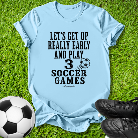 A light blue graphic t-shirt laid flat on green grass, featuring a black printed slogan about soccer. A soccer ball and black cleats are placed around it as props.