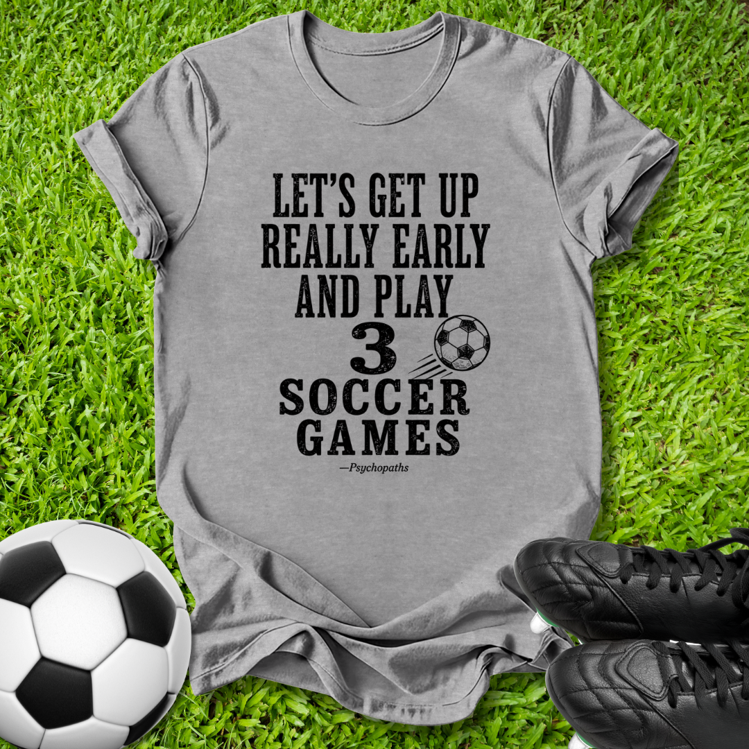 A heather gray graphic t-shirt laid flat on green grass, featuring the black printed text 'LET'S GET UP REALLY EARLY AND PLAY 3 SOCCER GAMES' with a soccer ball graphic. A soccer ball and black cleats are styled around the shirt.