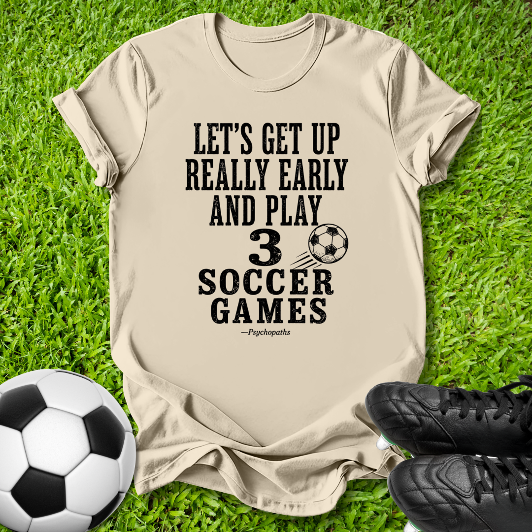 A beige graphic t-shirt laid flat on green grass, featuring the black printed text 'LET'S GET UP REALLY EARLY AND PLAY 3 SOCCER GAMES' with a soccer ball graphic and the attribution '—Psychopaths'. A black-and-white soccer ball is on the bottom left, and black soccer cleats are on the bottom right.