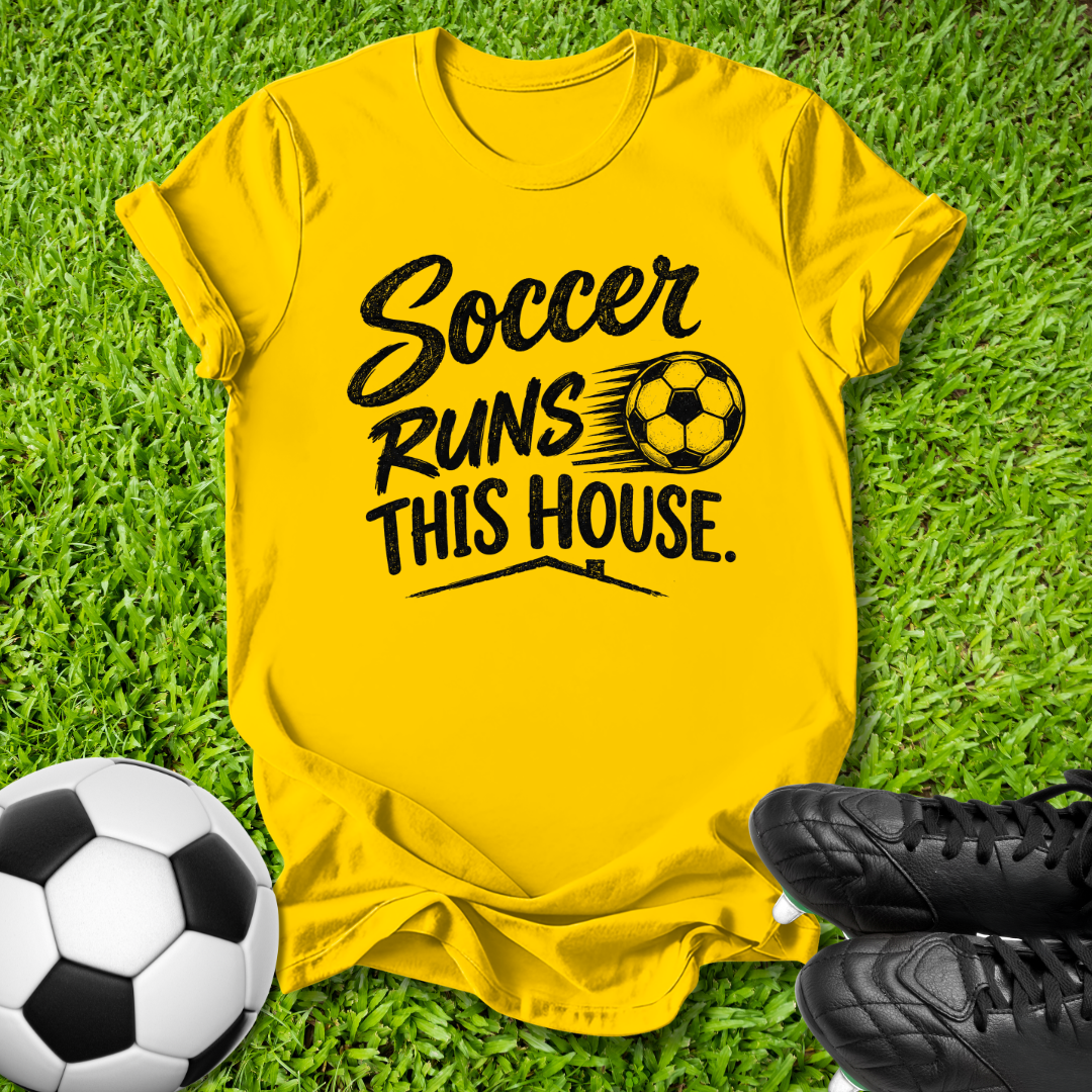 A bright yellow short-sleeve t-shirt laid flat on green grass, printed with the phrase 'Soccer Runs This House.' in black script and block lettering alongside a soccer ball graphic.