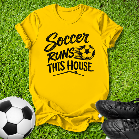 A bright yellow short-sleeve t-shirt laid flat on green grass, printed with the phrase 'Soccer Runs This House.' in black script and block lettering alongside a soccer ball graphic.