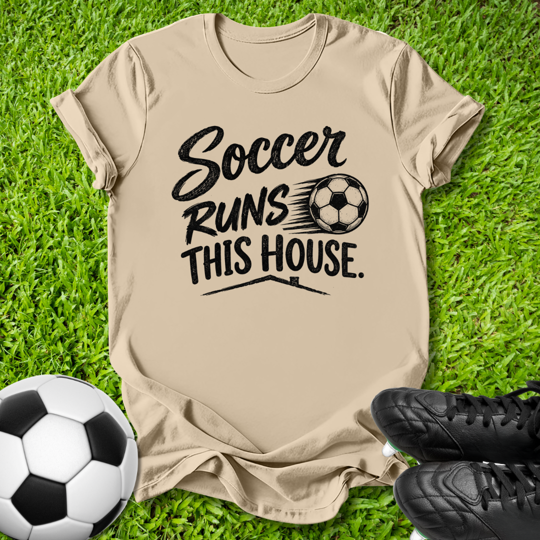 A beige graphic t-shirt laid flat on green grass, featuring the black text 'Soccer Runs This House.' alongside a soccer ball graphic.