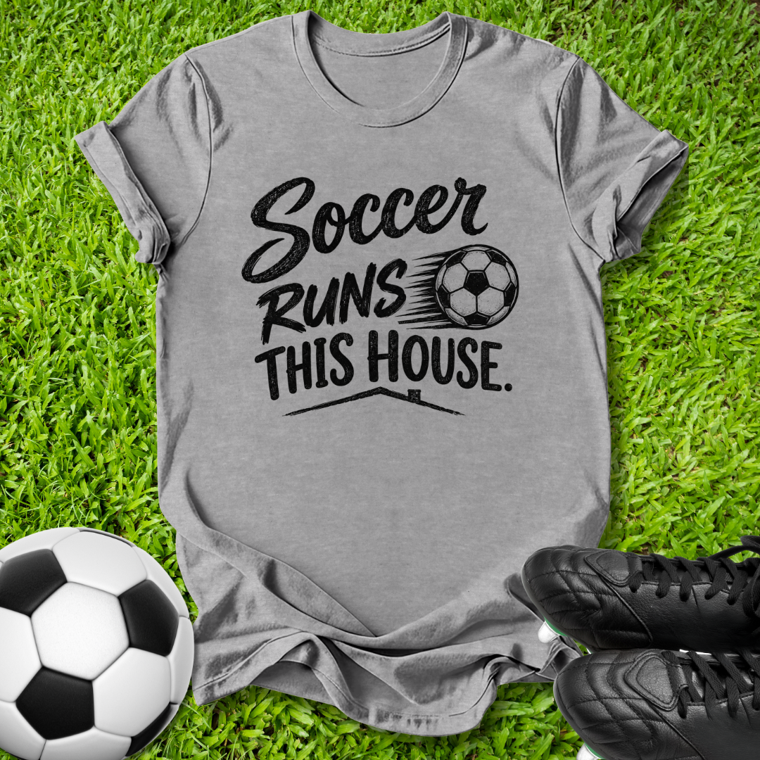 A heather gray short-sleeve t-shirt with a black graphic reading 'Soccer Runs This House.' and a soccer ball illustration, laid flat on green grass with a soccer ball and black cleats nearby.