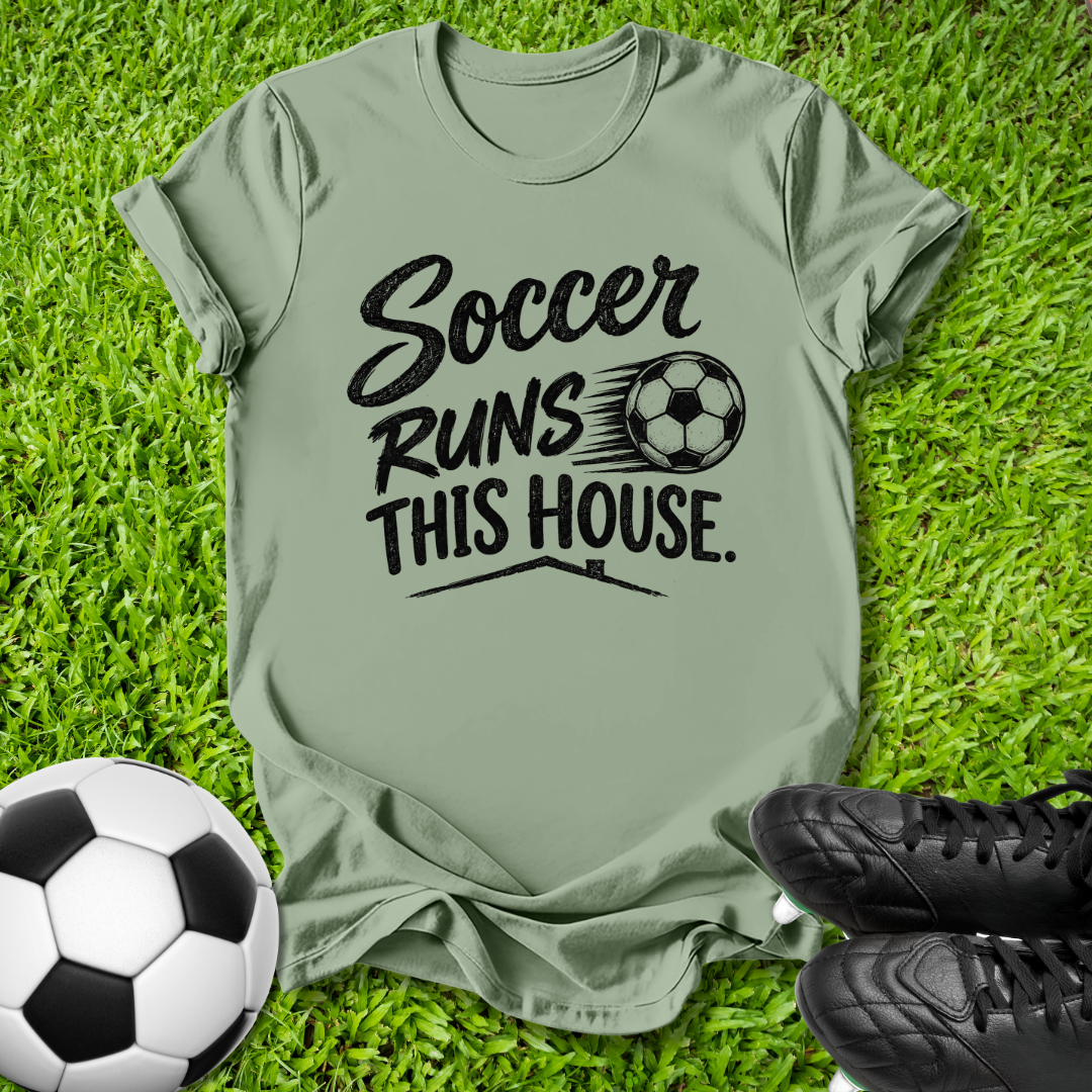 An olive green short-sleeve t-shirt with a black graphic reading 'Soccer Runs This House.' alongside a soccer ball illustration, laid flat on green grass with a soccer ball and black cleats nearby.