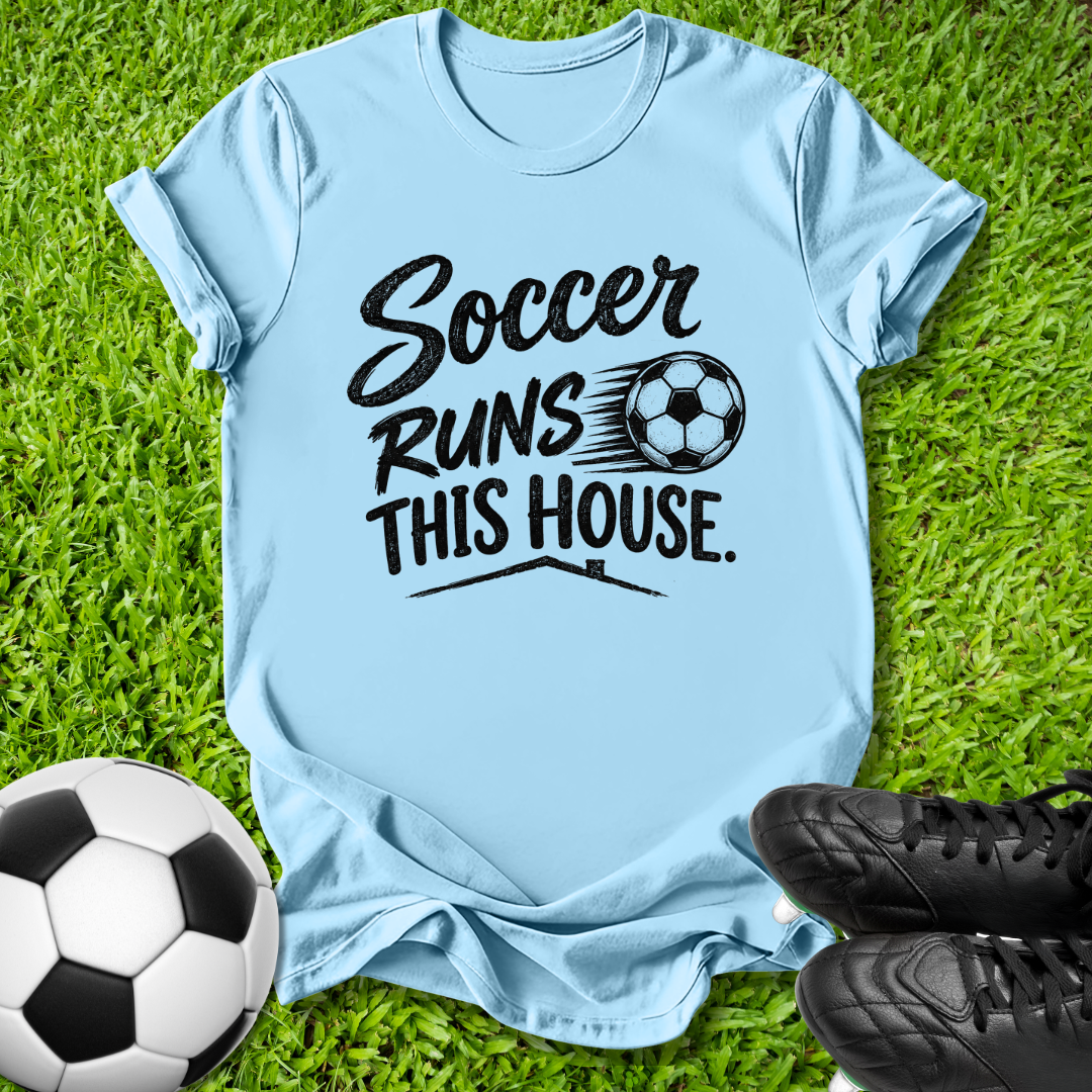 A light blue t-shirt with a black graphic reading 'Soccer Runs This House.' is laid flat on green grass, styled with a soccer ball and black cleats.