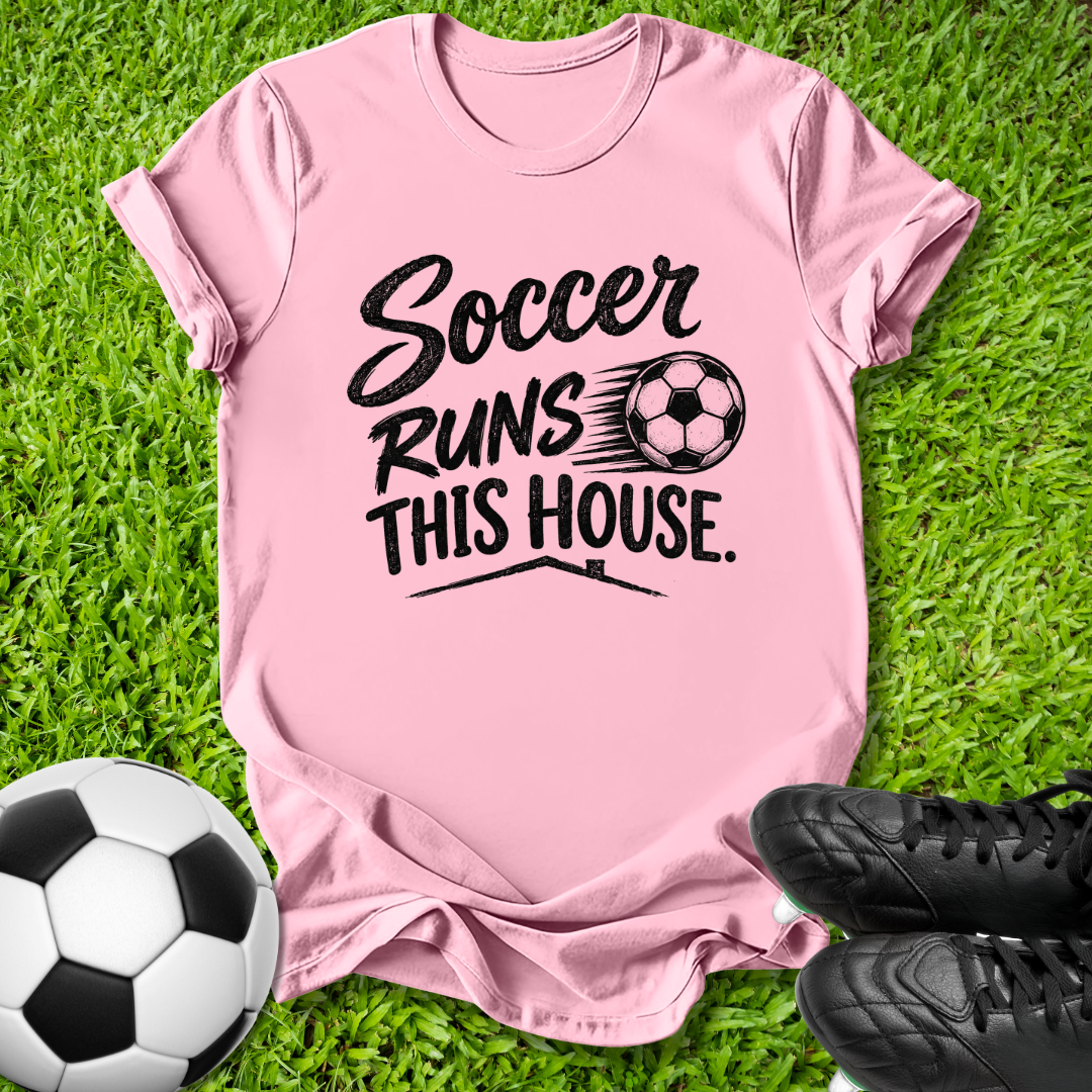 A pink short-sleeve t-shirt with a black graphic reading 'Soccer Runs This House.' and a soccer ball illustration, laid flat on green grass with a soccer ball and black cleats nearby.