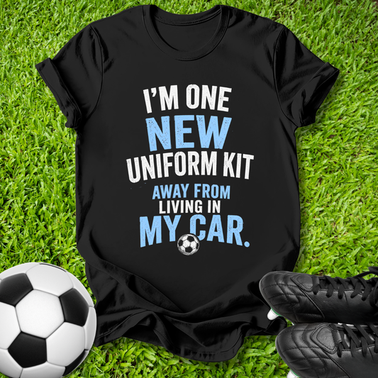 A black graphic t-shirt laid flat on green grass with soccer props, including a soccer ball and black cleats. The shirt features the text 'I'M ONE NEW UNIFORM KIT AWAY FROM LIVING IN MY CAR.