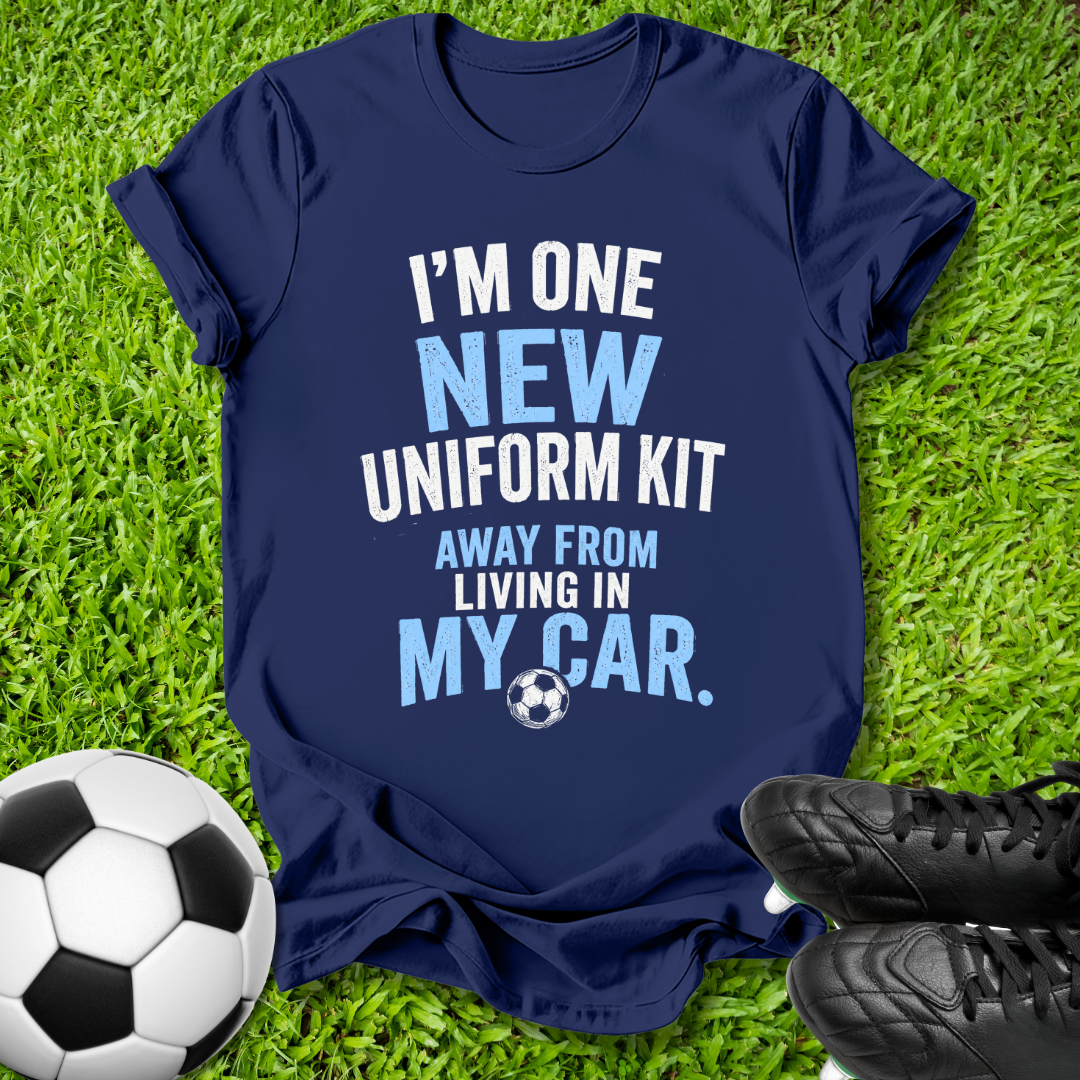 A navy blue graphic t-shirt laid flat on green grass, with a soccer ball on the left and black cleats on the right. The shirt features a white and light blue text graphic that reads, 'I'M ONE NEW UNIFORM KIT AWAY FROM LIVING IN MY CAR.