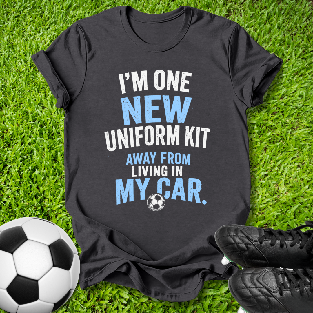 A black graphic t-shirt laid flat on green grass with a soccer ball and black cleats as props. The shirt features the text 'I'M ONE NEW UNIFORM KIT AWAY FROM LIVING IN MY CAR.
