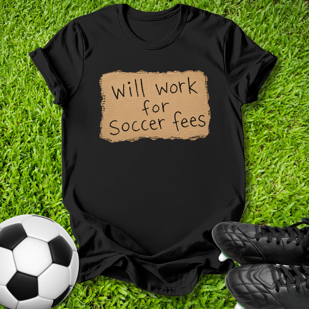 A black short-sleeve t-shirt laid flat on green grass, featuring a tan rectangular patch graphic with the handwritten text 'Will work for Soccer fees'. A black-and-white soccer ball is visible on the bottom left, and black cleats are visible on the bottom right.