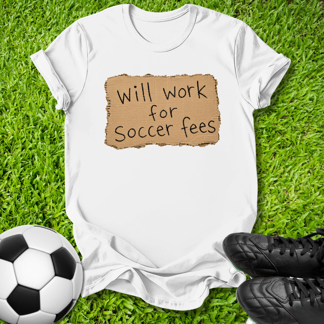 A white short-sleeve T-shirt laid flat on green grass, featuring a brown torn-paper style graphic with the text 'Will work for Soccer fees' in black. A soccer ball is at the lower left, and black cleats are at the lower right.