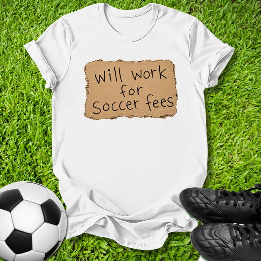 A white short-sleeve T-shirt laid flat on green grass, featuring a brown torn-paper style graphic with the text 'Will work for Soccer fees' in black. A soccer ball is at the lower left, and black cleats are at the lower right.