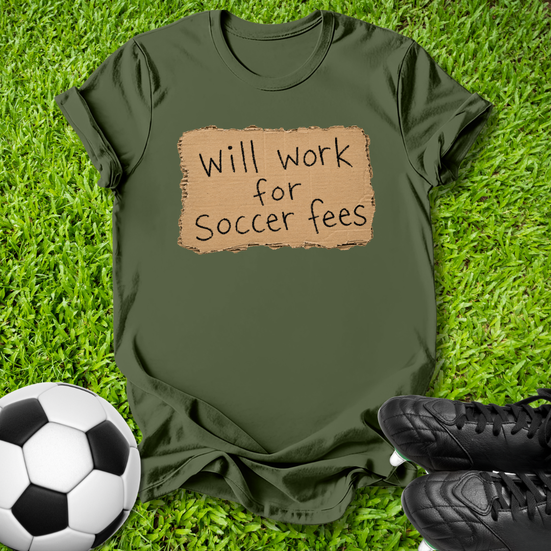 An olive green t-shirt with a cardboard-texture graphic that reads 'Will work for Soccer fees' in black handwritten text, laid flat on green grass with a soccer ball and black cleats nearby.