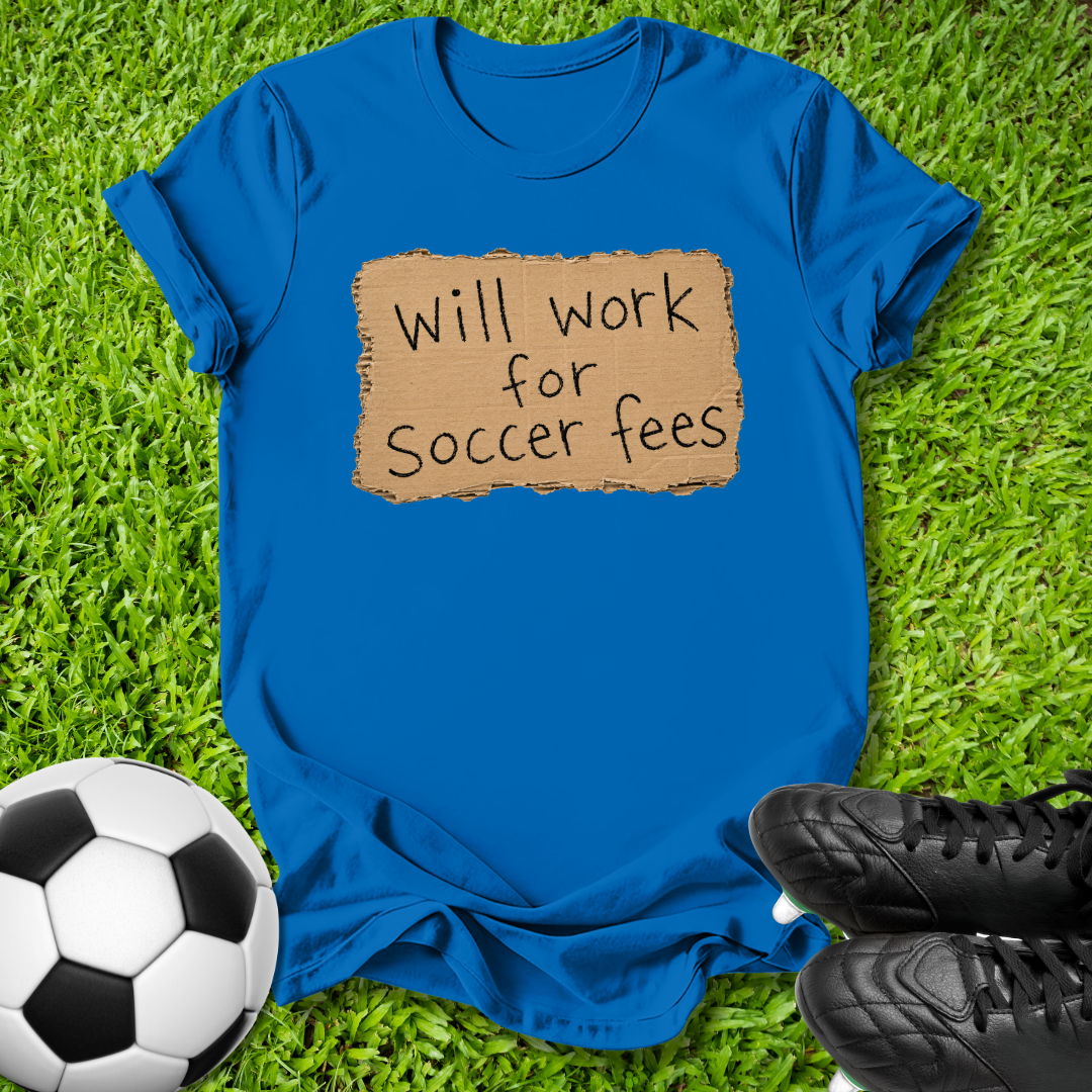 A bright blue short-sleeve t-shirt laid flat on green grass, featuring a tan rectangular patch with the handwritten text 'Will work for Soccer fees' in black. A soccer ball is visible on the lower left, and black cleats are on the lower right.