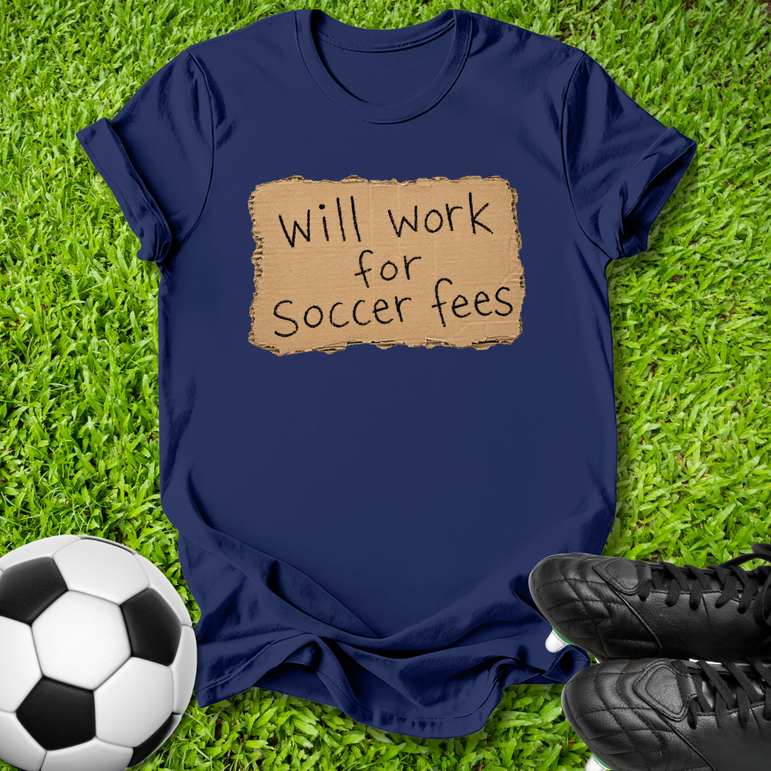 A navy blue short-sleeve t-shirt laid flat on green grass, featuring a tan rectangular graphic with the handwritten text 'Will work for Soccer fees'. A black-and-white soccer ball is visible in the lower-left corner, and black soccer cleats are in the lower-right corner.