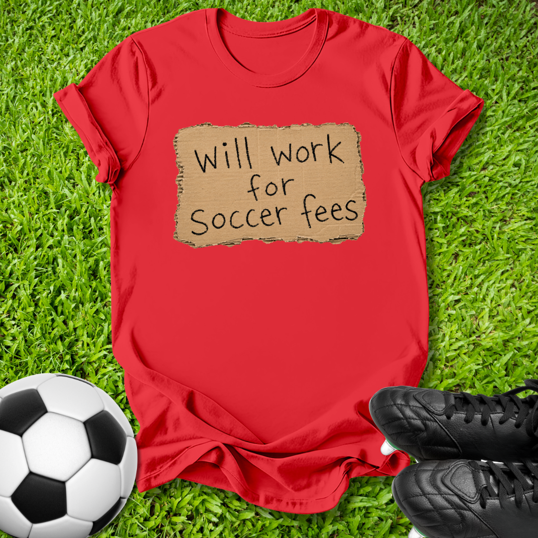 A red short-sleeve t-shirt laid flat on green grass, featuring a cardboard-texture graphic that reads 'Will work for Soccer fees' in black handwritten text. A black-and-white soccer ball is at the bottom left, and black soccer cleats are at the bottom right.