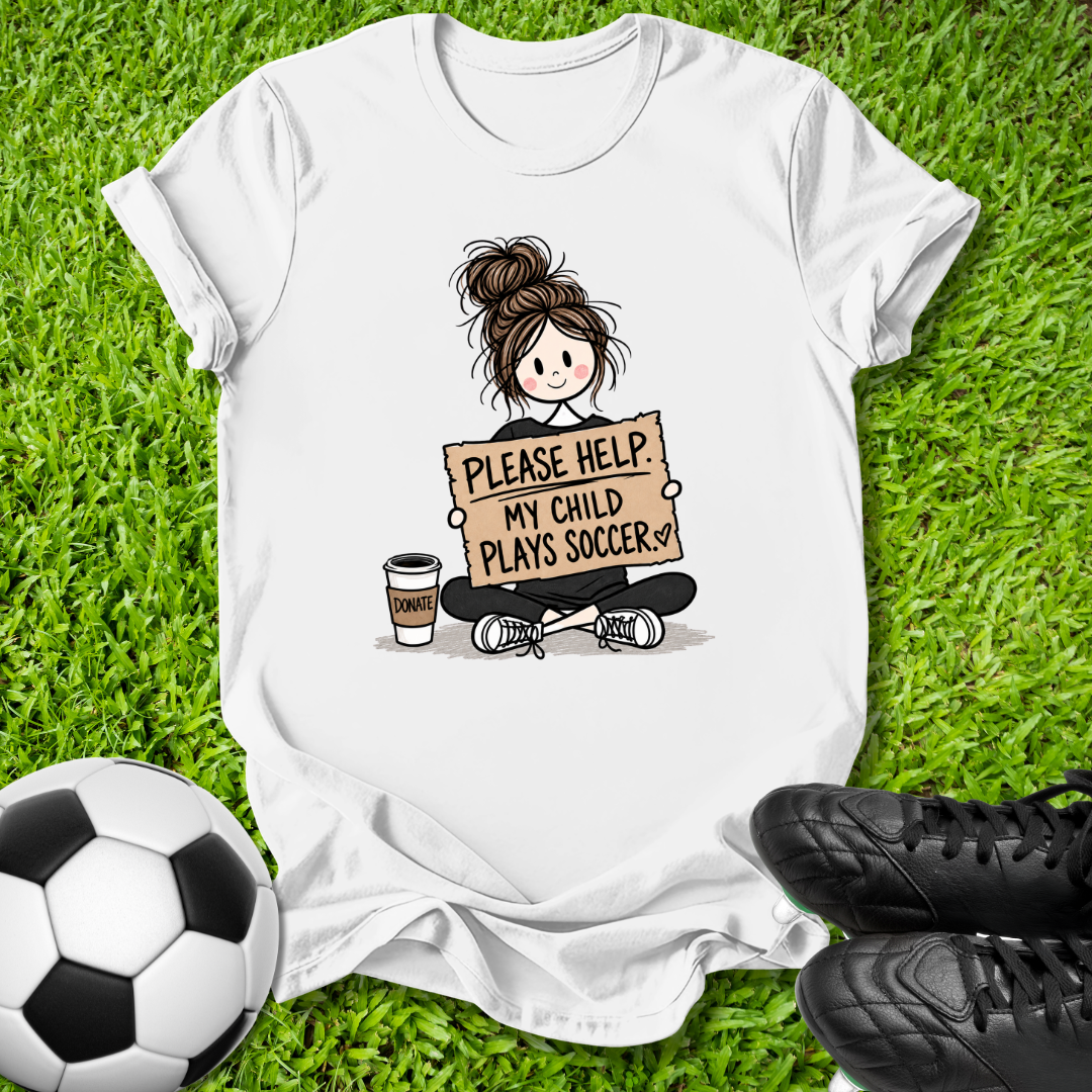 A white graphic t-shirt laid flat on green grass, featuring a cartoon illustration of a girl sitting cross-legged and holding a sign that reads 'PLEASE HELP MY CHILD PLAYS SOCCER'. A soccer ball is visible on the bottom left and black soccer cleats are visible on the bottom right.