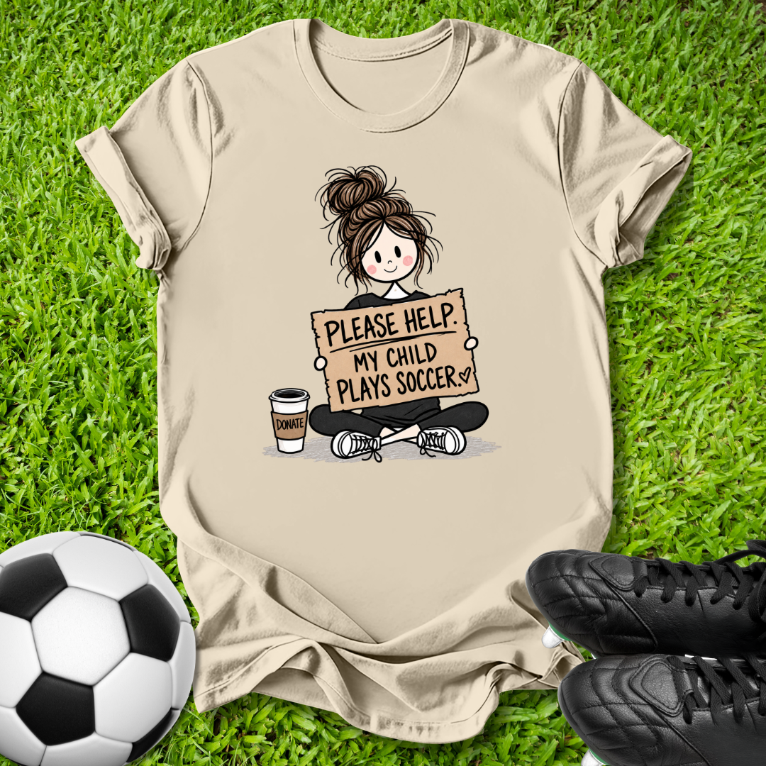 A beige graphic t-shirt laid flat on green grass, featuring a cartoon illustration of a girl sitting cross-legged holding a sign that reads 'PLEASE HELP MY CHILD PLAYS SOCCER'. A soccer ball is on the bottom left, and black soccer cleats are on the bottom right.