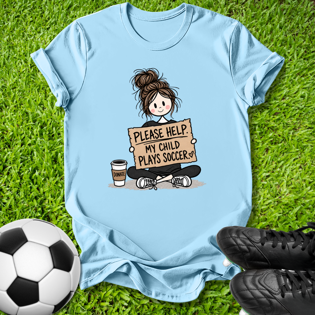 A light blue graphic t-shirt laid flat on green grass, featuring a cartoon illustration of a girl holding a sign that reads 'PLEASE HELP MY CHILD PLAYS SOCCER'. A soccer ball is visible on the bottom left and black soccer cleats are on the bottom right.