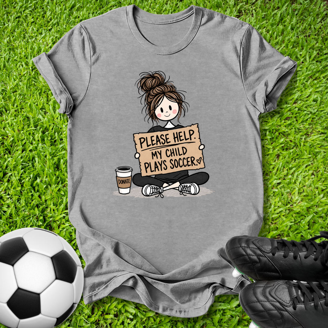 A heather gray graphic t-shirt laid flat on green grass, featuring a cartoon illustration of a soccer player holding a sign that reads 'PLEASE HELP MY CHILD PLAYS SOCCER'. A soccer ball and black cleats are styled around the shirt.