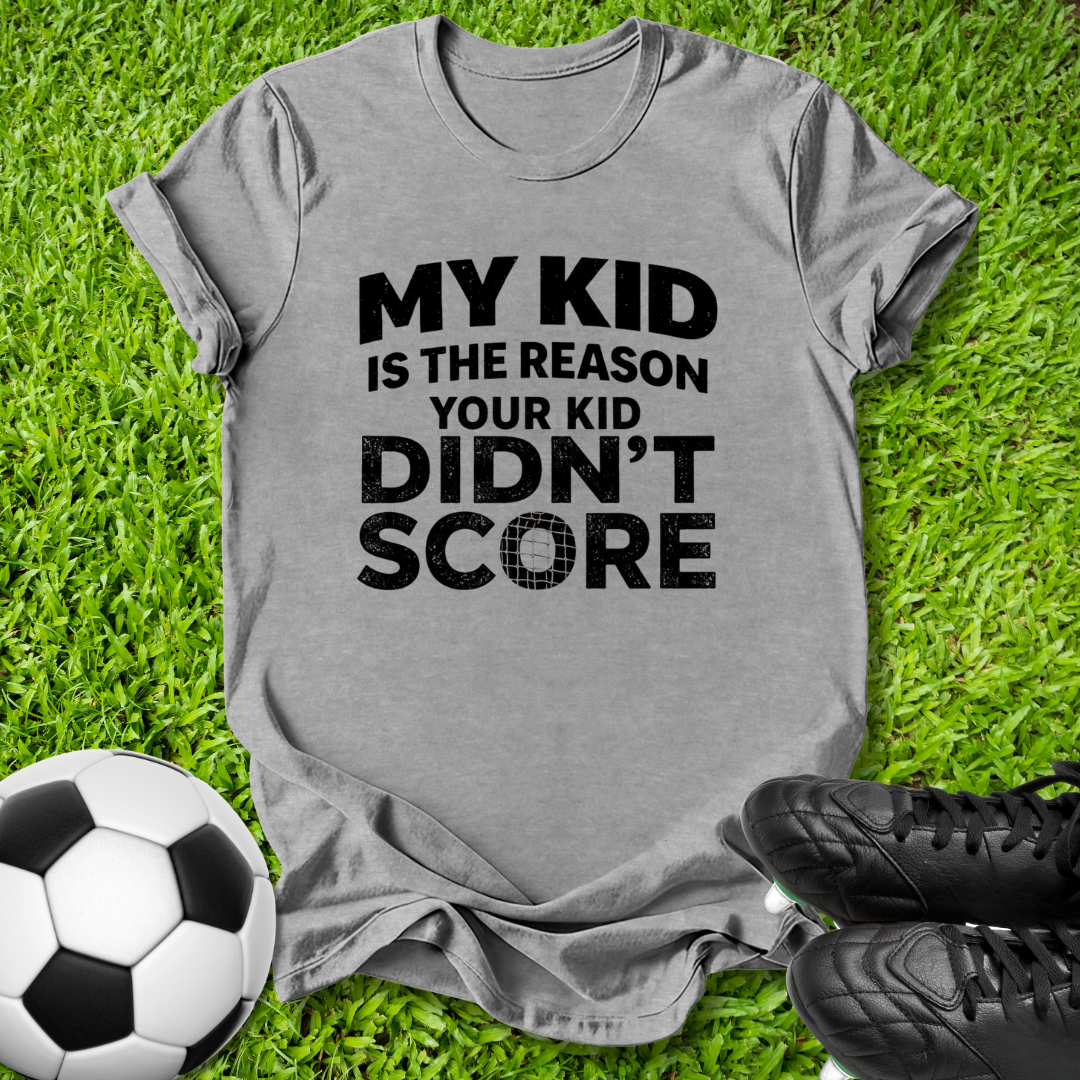 A heather gray short-sleeve t-shirt with black text reading 'MY KID IS THE REASON YOUR KID DIDN'T SCORE', laid flat on green grass with a soccer ball and black cleats nearby.