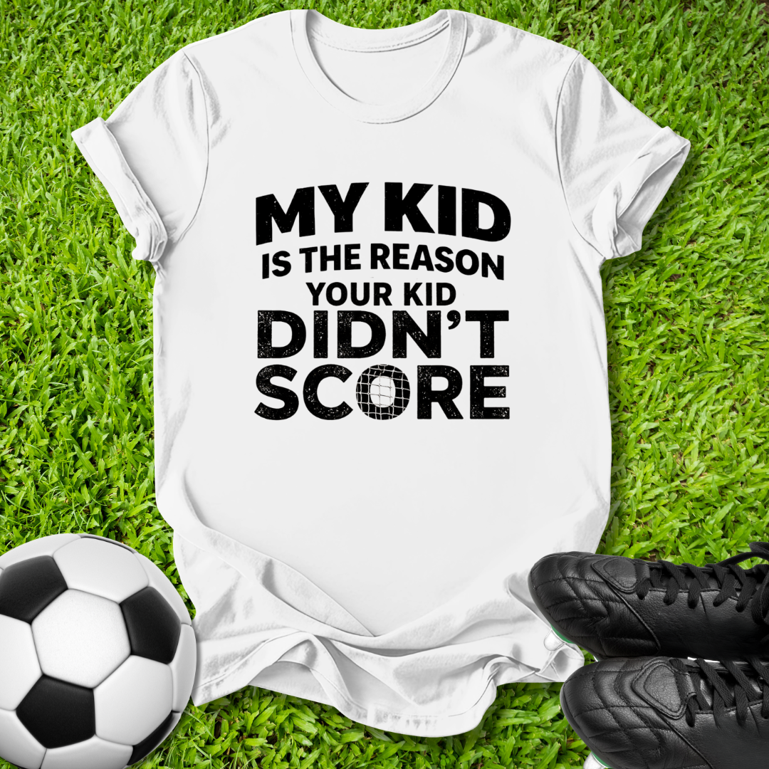 A white short-sleeve graphic T-shirt laid flat on green grass, with a soccer ball on the left and black cleats on the right. The shirt has bold black text reading, 'MY KID IS THE REASON YOUR KID DIDN'T SCORE.