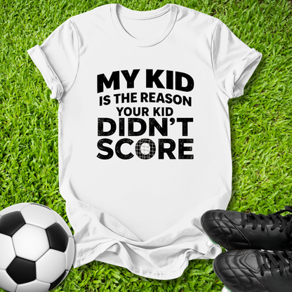 A white short-sleeve graphic T-shirt laid flat on green grass, with a soccer ball on the left and black cleats on the right. The shirt has bold black text reading, 'MY KID IS THE REASON YOUR KID DIDN'T SCORE.