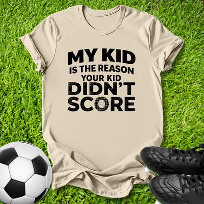 A beige graphic t-shirt laid flat on green grass, featuring the black text 'MY KID IS THE REASON YOUR KID DIDN'T SCORE'. A soccer ball is on the left and black cleats are on the right.