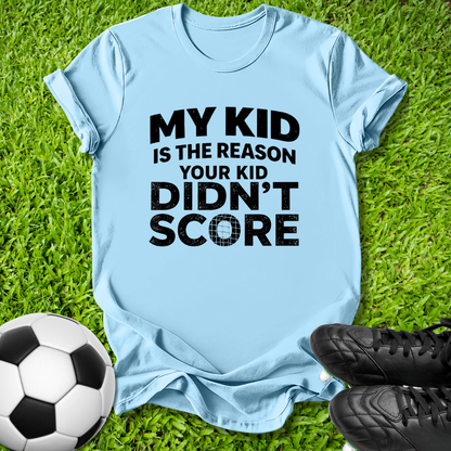 A light blue graphic t-shirt laid flat on green grass, featuring the text 'MY KID IS THE REASON YOUR KID DIDN'T SCORE' in bold black lettering. A soccer ball and black cleats are styled around the shirt.
