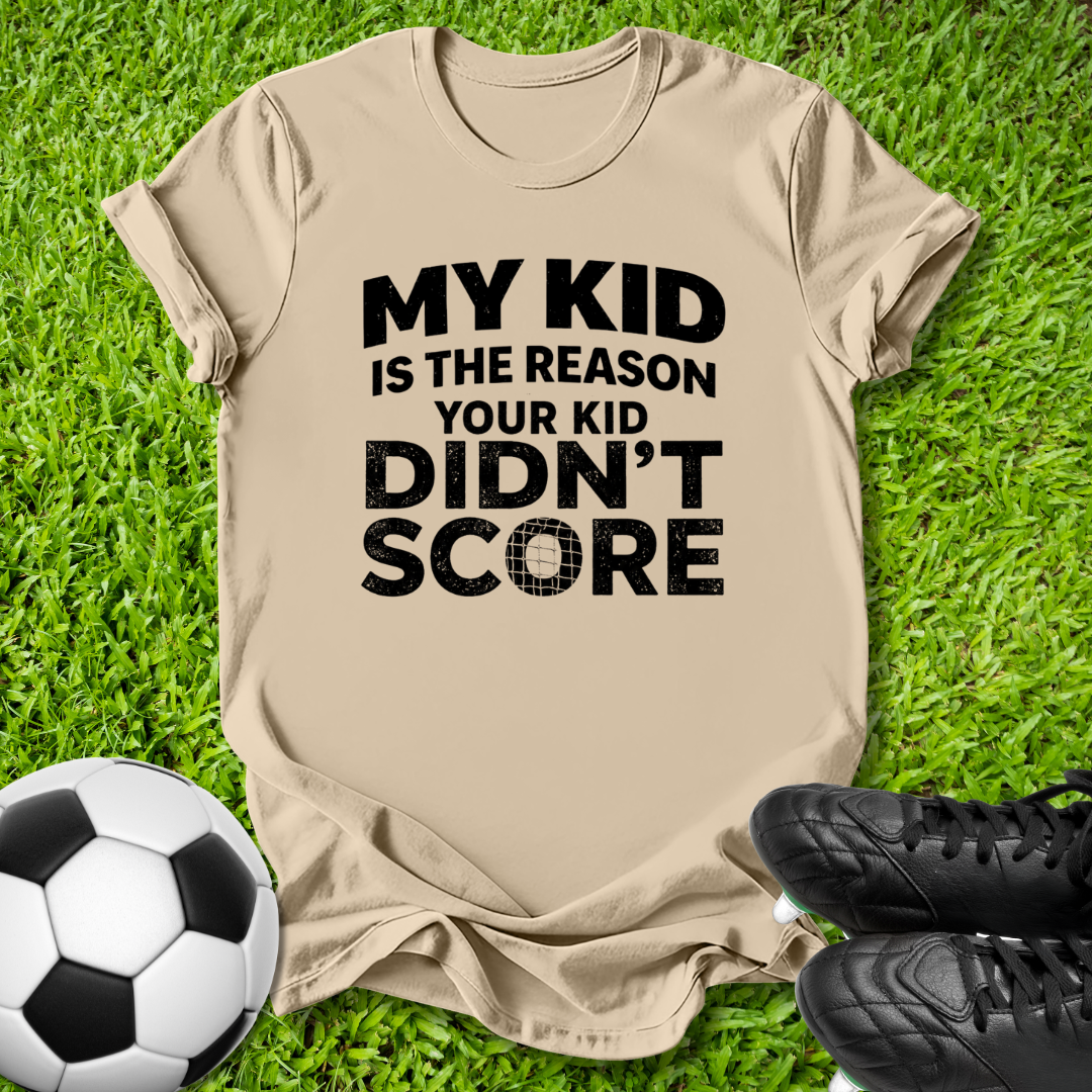 A beige graphic t-shirt laid flat on green grass, featuring the bold black text 'MY KID IS THE REASON YOUR KID DIDN'T SCORE'. A soccer ball is visible in the bottom-left corner and a pair of black soccer cleats is in the bottom-right, creating a sports-themed flat-lay composition.
