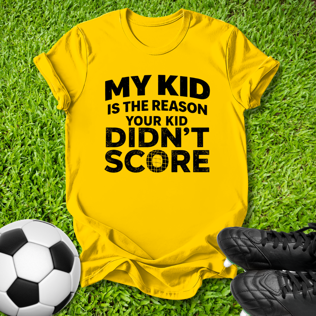 A bright yellow graphic t-shirt laid flat on green grass, featuring the bold black text 'MY KID IS THE REASON YOUR KID DIDN'T SCORE'. A soccer ball and black cleats are placed around it as thematic props.