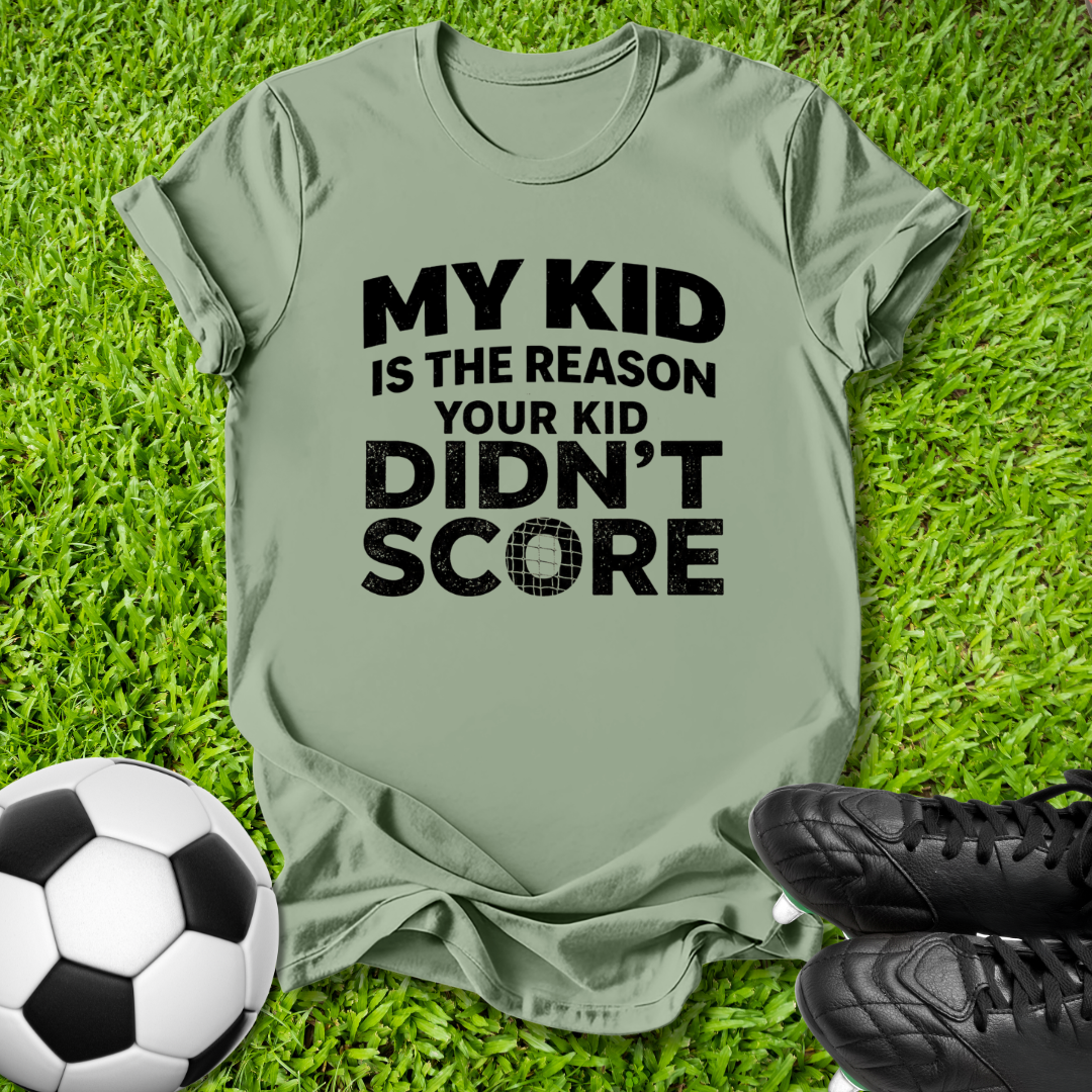 An olive green t-shirt with black text reading 'MY KID IS THE REASON YOUR KID DIDN'T SCORE' laid flat on green grass, styled with a soccer ball and black cleats.