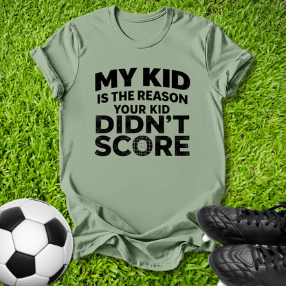 An olive green t-shirt with black text reading 'MY KID IS THE REASON YOUR KID DIDN'T SCORE' laid flat on green grass, styled with a soccer ball and black cleats.