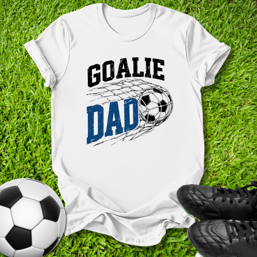 A white short-sleeve graphic T-shirt laid flat on green grass, featuring the words 'GOALIE DAD' with a soccer ball and net design. A soccer ball and black cleats are placed around it as props.