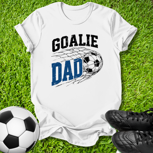 A white short-sleeve graphic T-shirt laid flat on green grass, featuring the words 'GOALIE DAD' with a soccer ball and net design. A soccer ball and black cleats are placed around it as props.