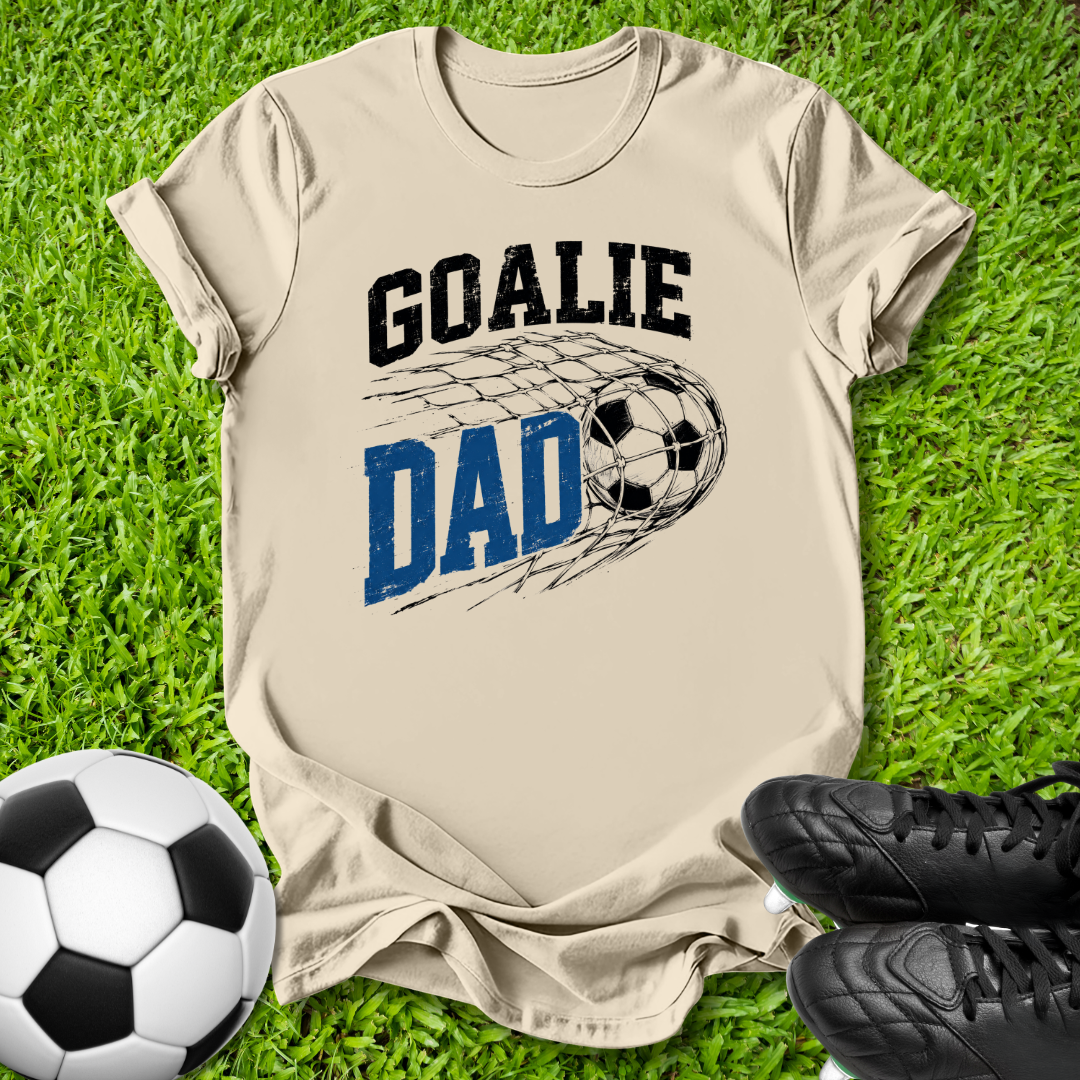 A beige graphic t-shirt laid flat on green grass, featuring the words 'GOALIE DAD' with a soccer ball and net design. A soccer ball and black cleats are placed around it as props.
