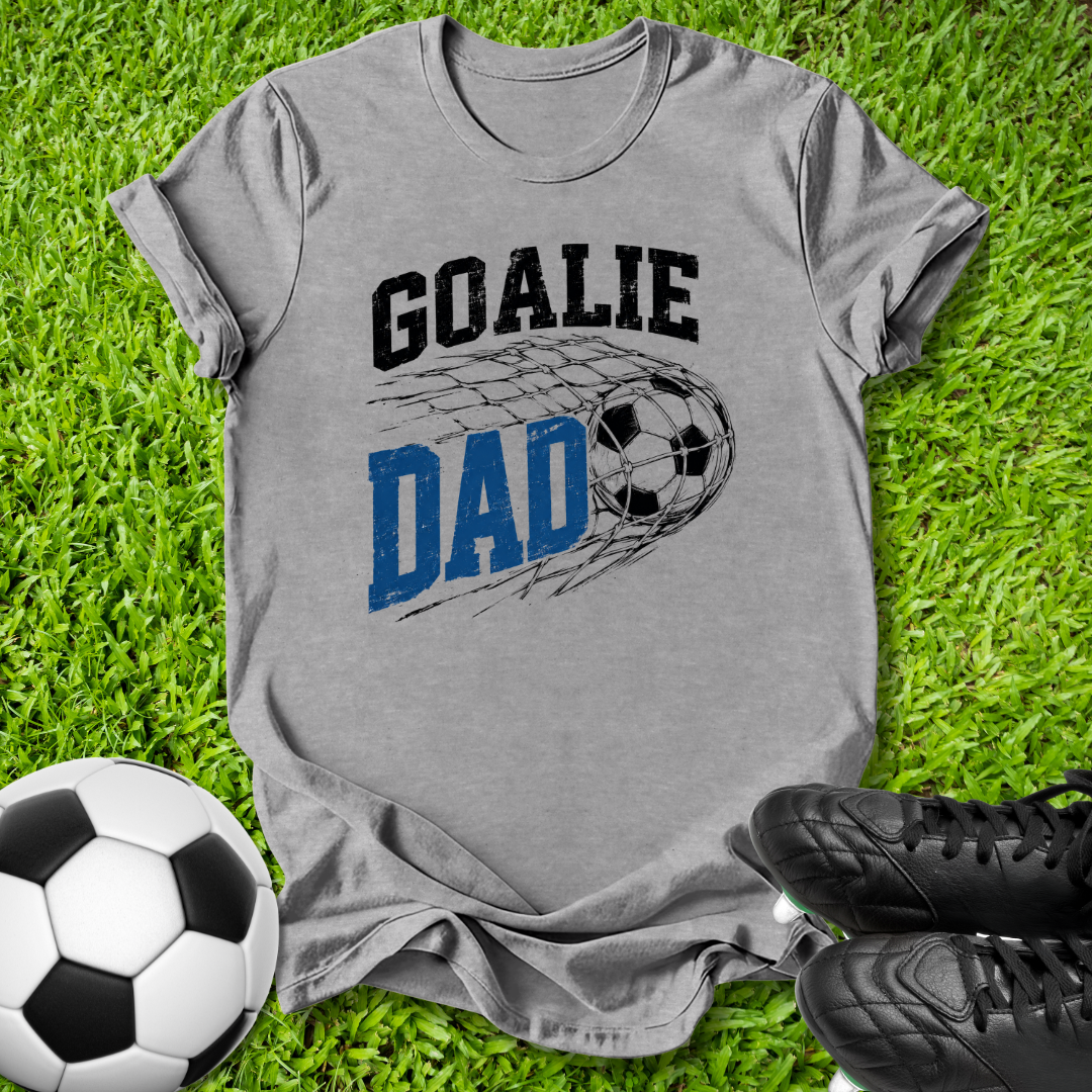 A heather gray graphic t-shirt laid flat on green grass, featuring the words 'GOALIE DAD' with a soccer ball and net design. A soccer ball and black cleats are styled around the shirt.