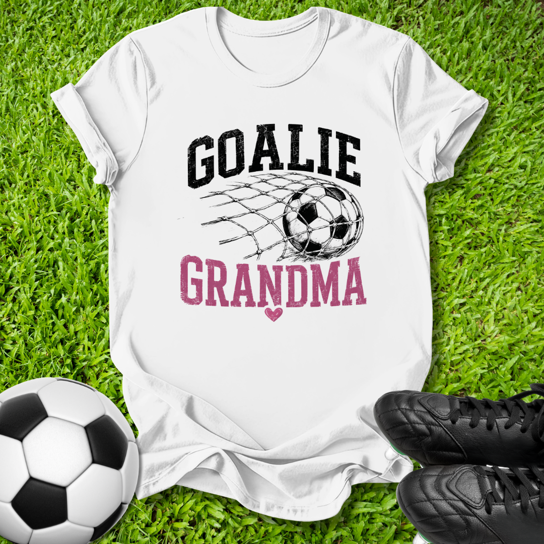 A white short-sleeve graphic T-shirt laid flat on green grass, with a soccer ball net design and the words 'GOALIE GRANDMA' in black and pink. A soccer ball and black cleats are placed around it as props.