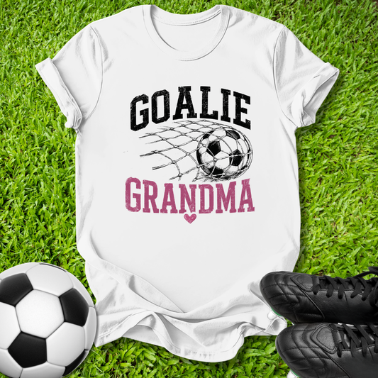 A white short-sleeve graphic T-shirt laid flat on green grass, with a soccer ball net design and the words 'GOALIE GRANDMA' in black and pink. A soccer ball and black cleats are placed around it as props.