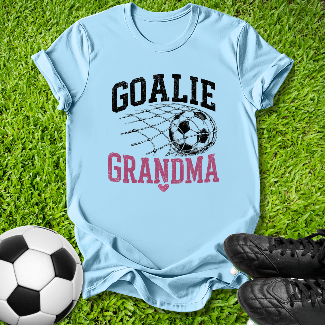 A light blue graphic t-shirt laid flat on green grass, featuring the words 'GOALIE GRANDMA' with a soccer ball and net design. A soccer ball and black cleats are placed around it as props.