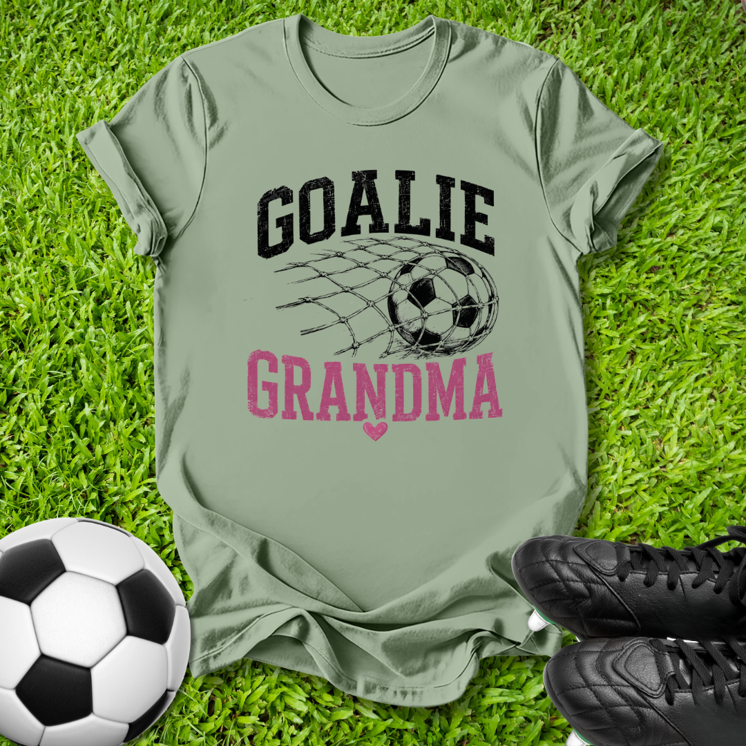 An olive green graphic t-shirt laid flat on green grass, featuring a black soccer net and ball design with the words 'GOALIE GRANDMA' in black and pink. A soccer ball and black cleats are placed around it as props.